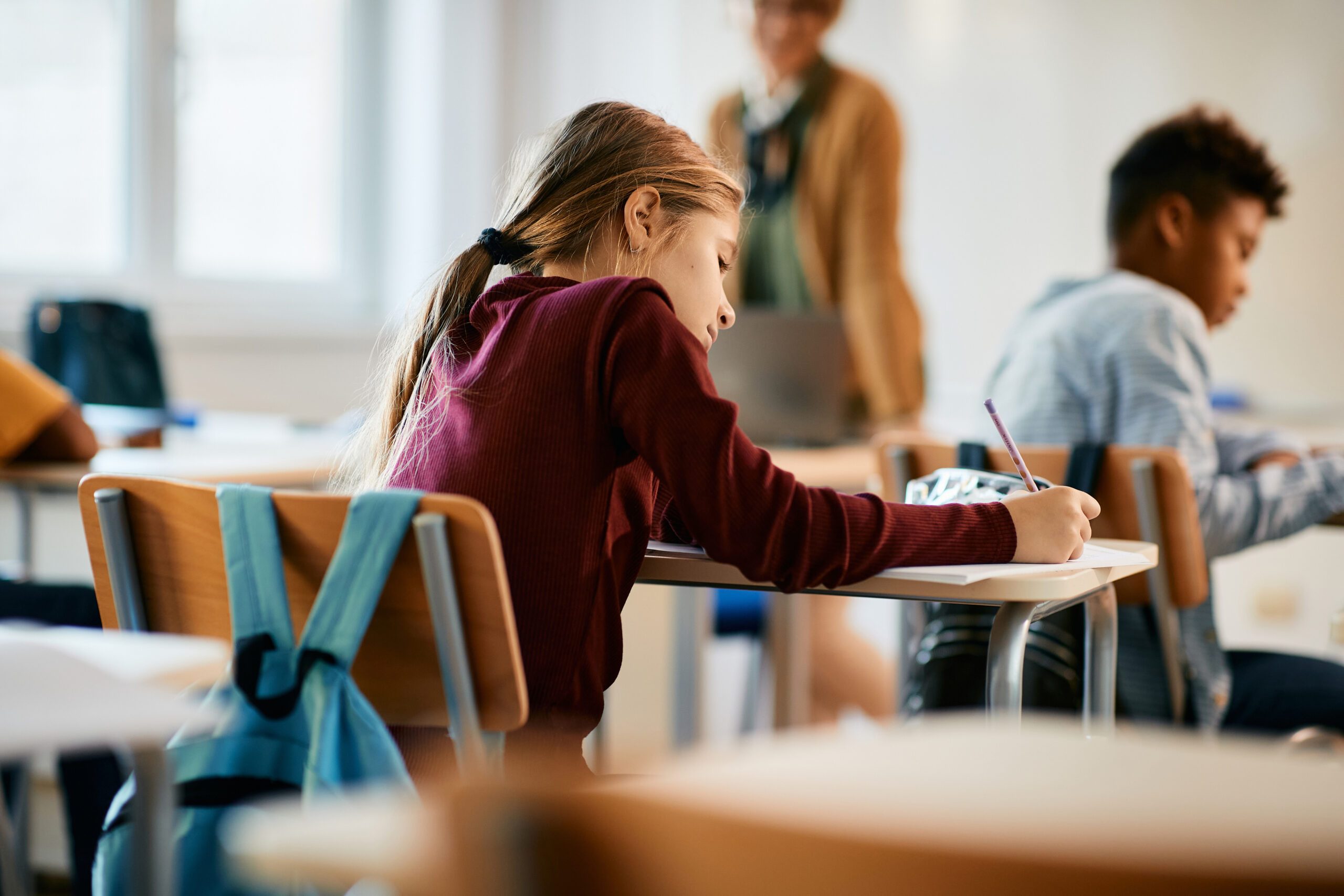 Bakre vy av skolflicka som sitter och skriver vid sin skolbänk under lektionstid.