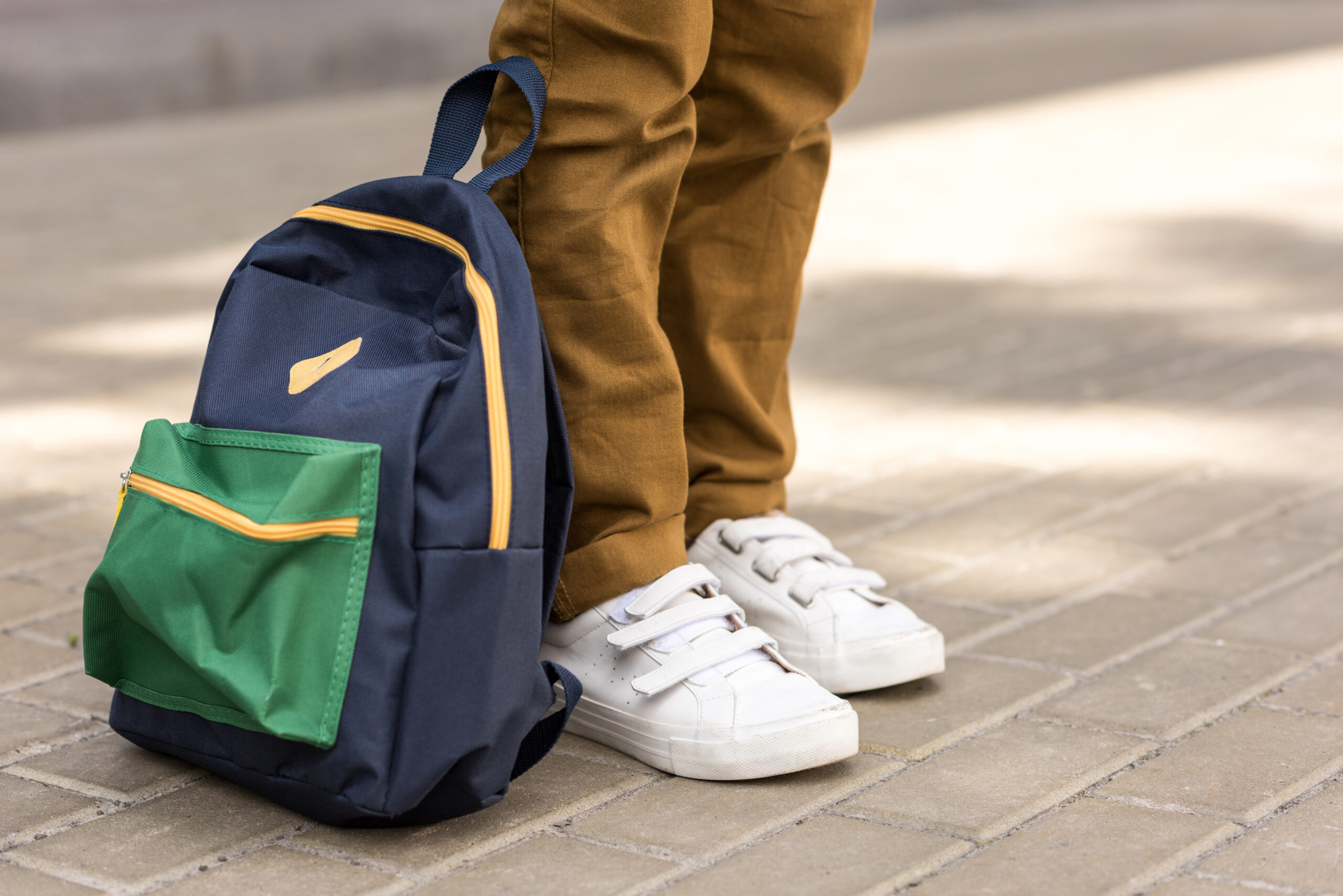 Närbild av en skolelevs skor som har sin ryggsäck stående bredvid sig på marken.
